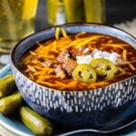 A large blue bowl filled with Texas chili topped with cheese, onions, and jalapenos next to a glass of beer and a jar of pickles.