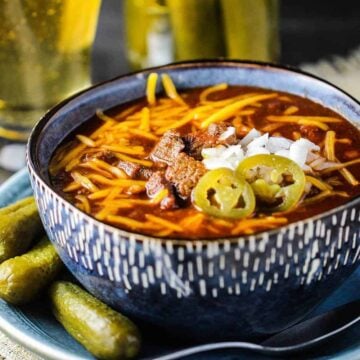 A large blue bowl filled with Texas chili topped with cheese, onions, and jalapenos next to a glass of beer and a jar of pickles.