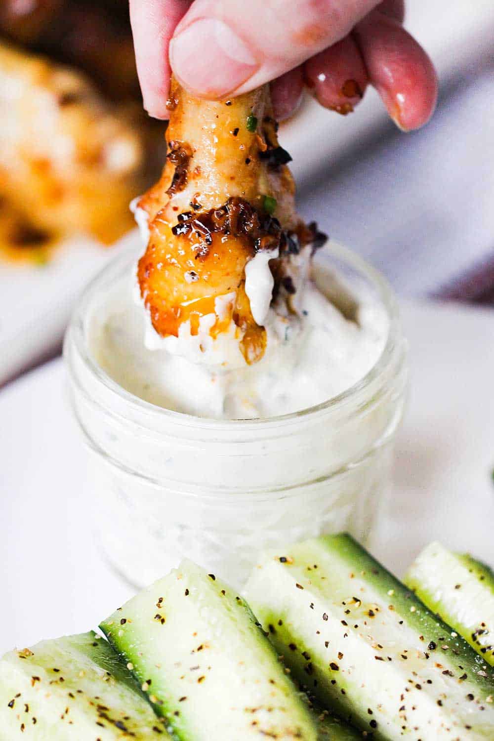 A hand plunging a baked hot wing into a jar of blue cheese dressing next to a plate of sliced cucumbers.