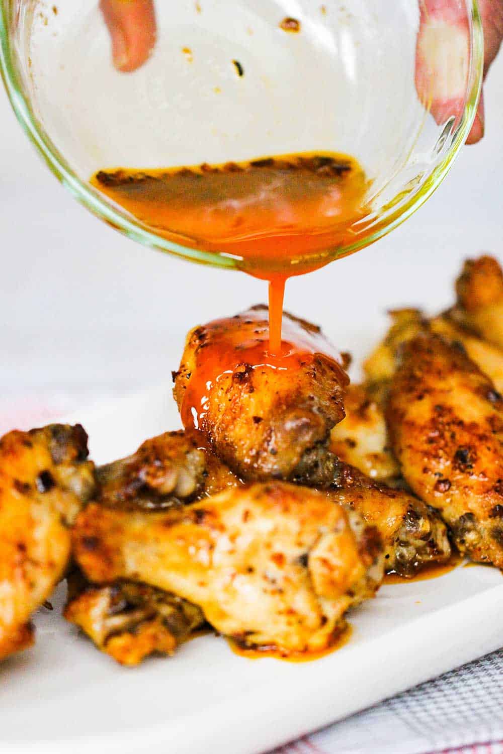 A hand pouring hot sauce from a small bowl over baked hot wings.