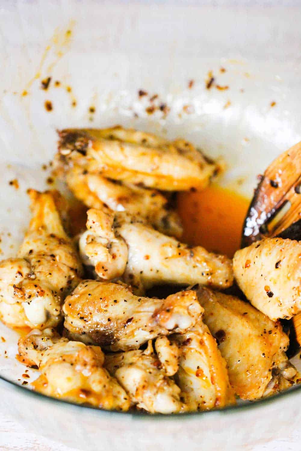 A glass bowl of hot wings that are being tossed with a wooden spoon.