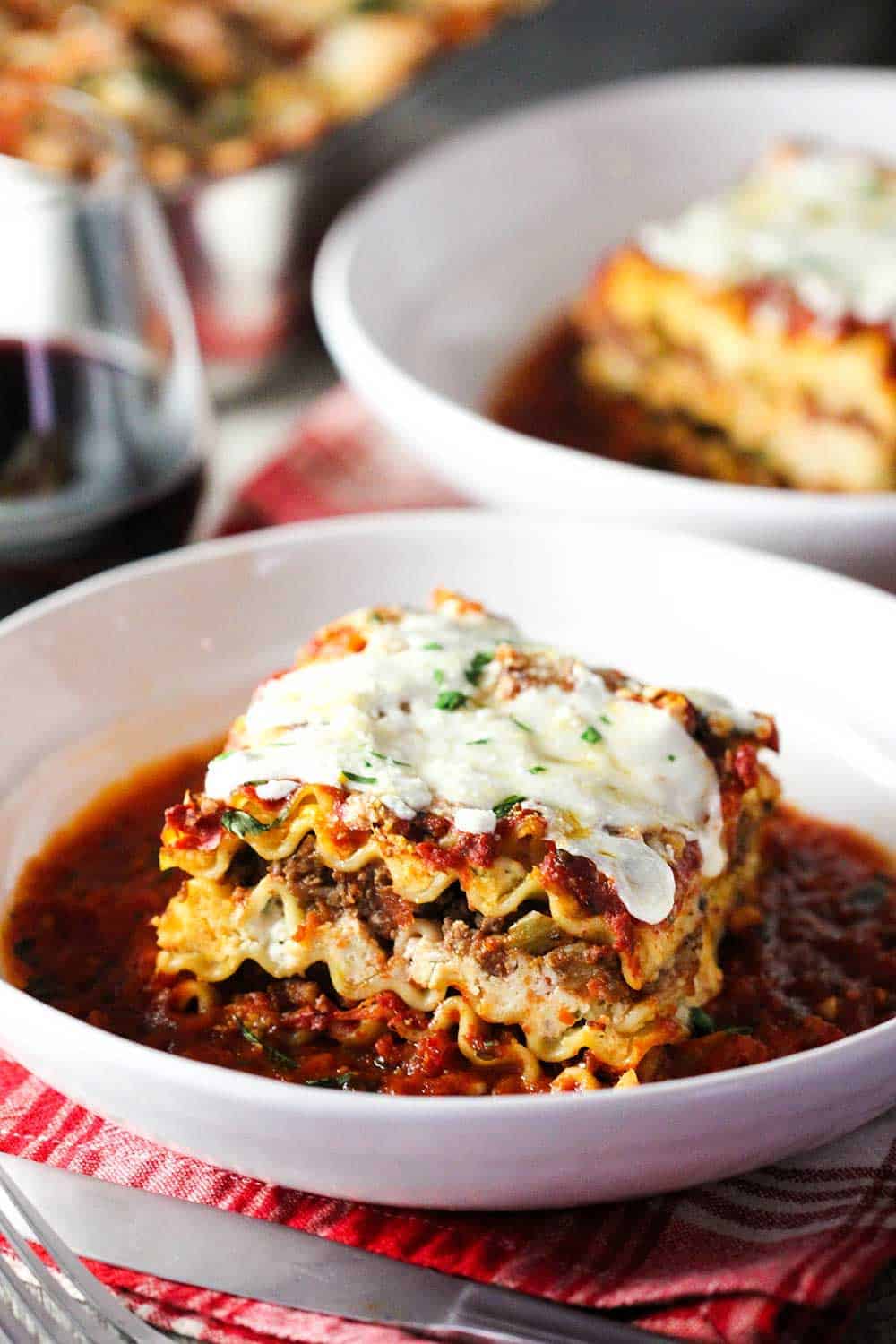 A white bowl holding classic meat lasagna with another bowl behind it next to a glass of red wine.