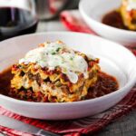 A white bowl of classic meat lasagna next to a glass of red wine.