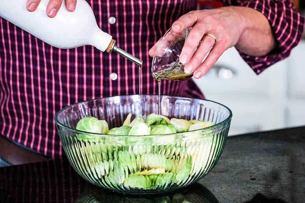 A person pouring bacon grease from a small dish in one hand, and pouring olive oil from a container with the other hand into a bowl of uncooked Brussels sprouts.