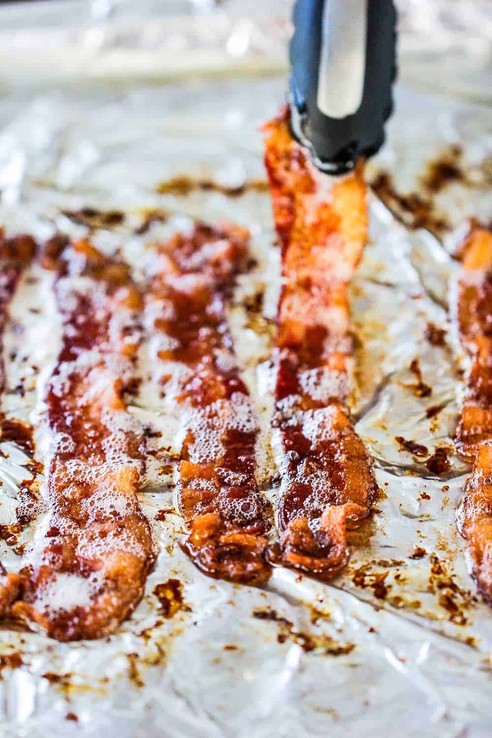 Cooked bacon slices on a baking sheet lined with foil with a pair of tongs picking one slice up.
