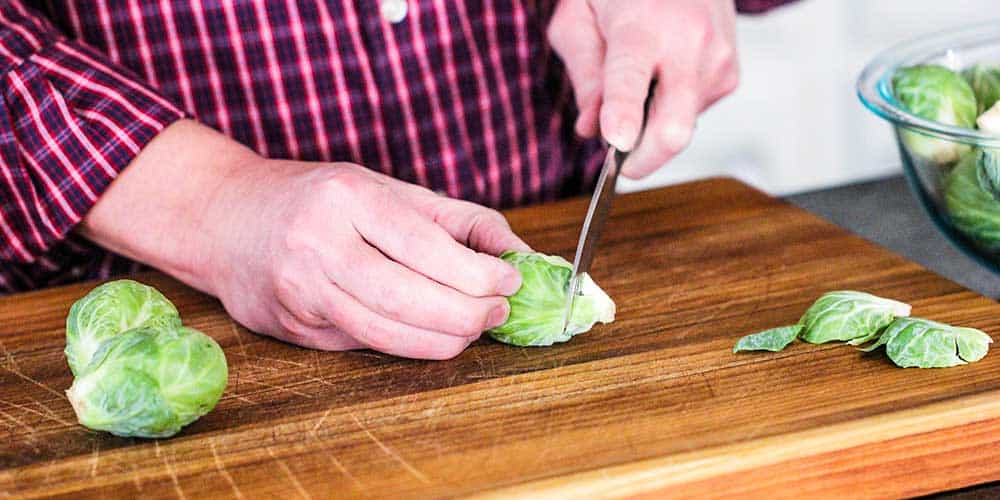 Two hands clipping the end off of a Brussels sprout on a wooden cutting board.