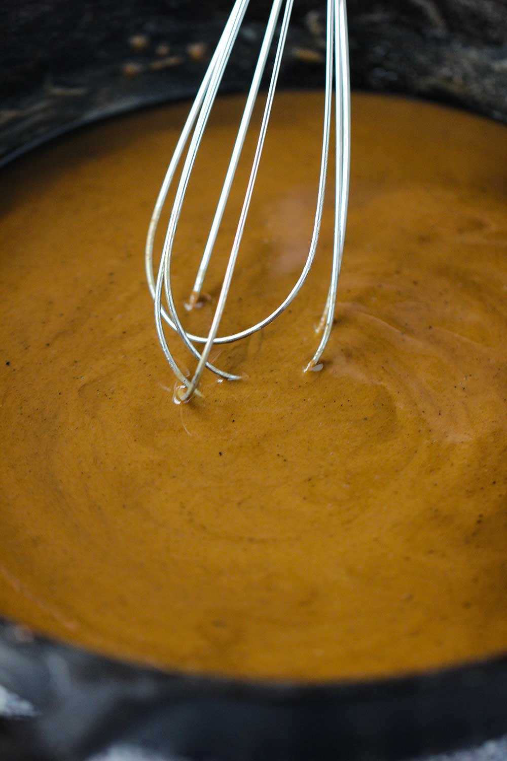 A brown roux being stirred with a whisk in a black cast-iron skillet.