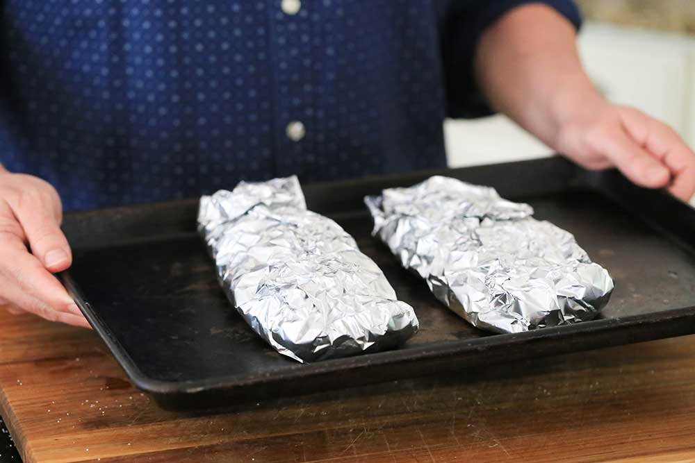 Two hands holding a baking sheet with two foil packets on it.