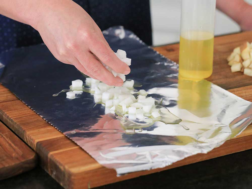 Two hands placing chopped onions on a rectangle of foil with a bottle of oil next to it.