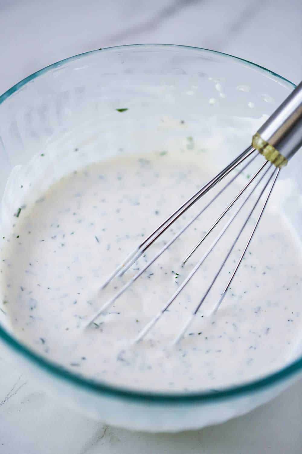 A glass bowl filled with ranch dressing with a wire whisk in it.