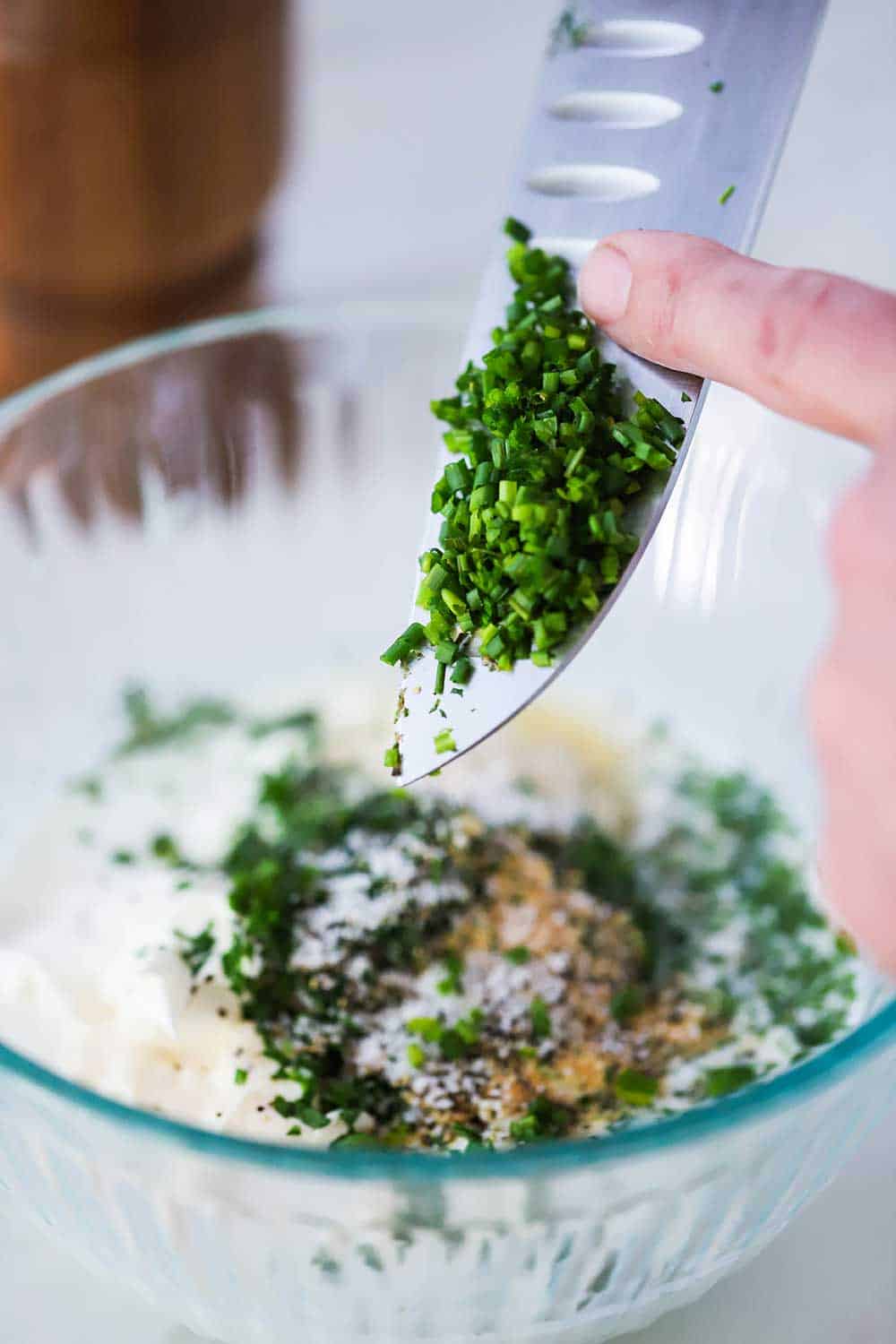 A hand scraping chopped chives into a bowl of Ranch dressing ingredients.
