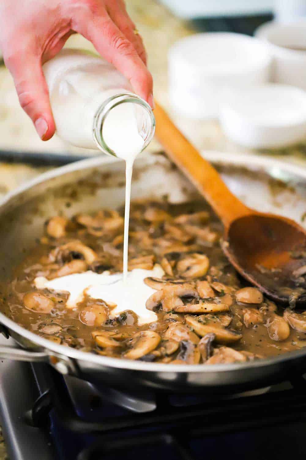A hand pouring cream into a skillet of sautéed mushrooms