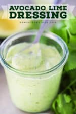 A small glass jar holding freshly made avocado lime dressing next to a small spoon and a bunch of fresh parsley and a cut avocado in the background.