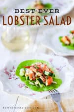 A mound of lobster salad with cucumber on a leaf of lettuce on a decorative plate next to a glass of white wine and a platter.
