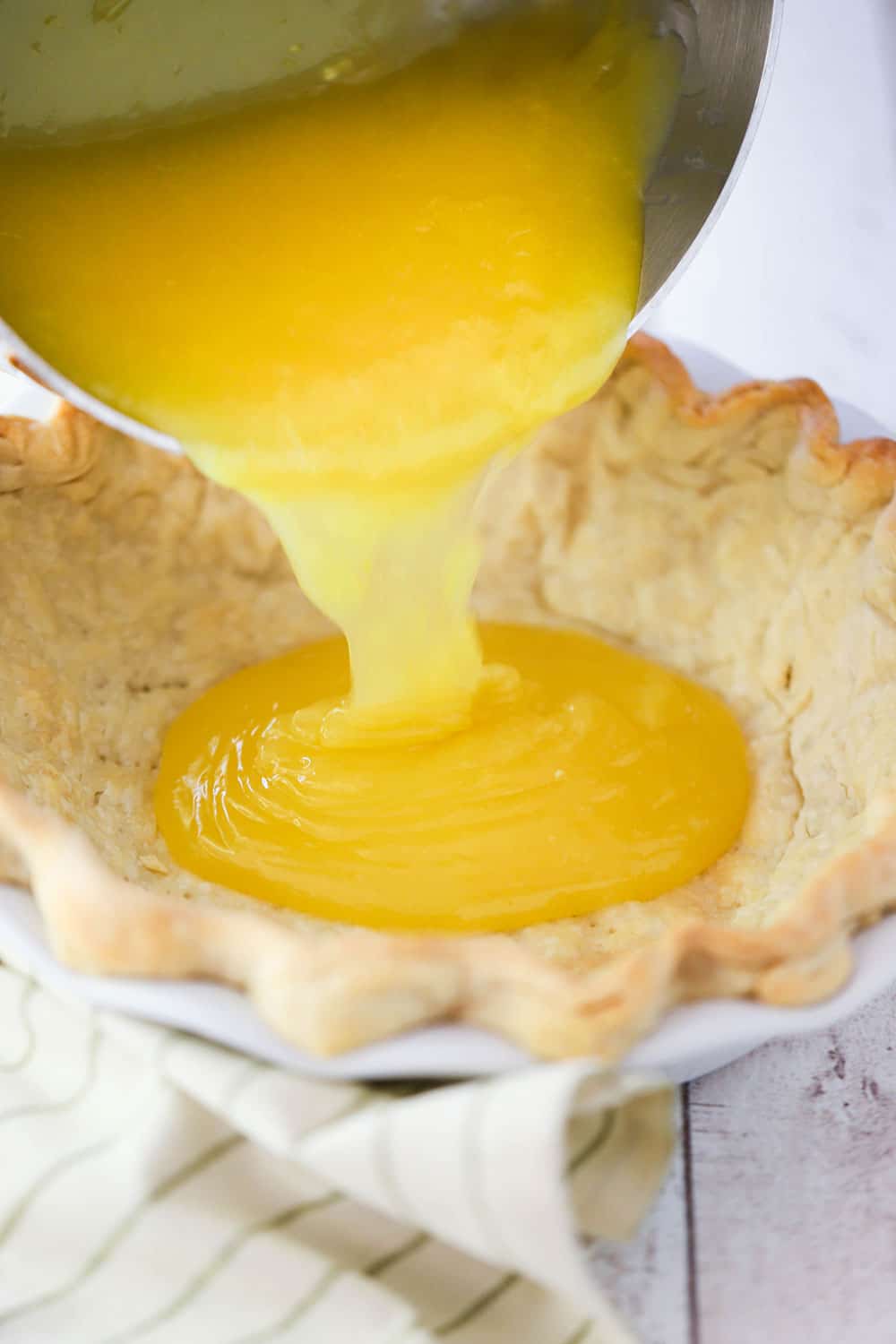 Lemon curd being poured from a metal bowl into a baked pie shell in a white pie dish next to a striped napkin.