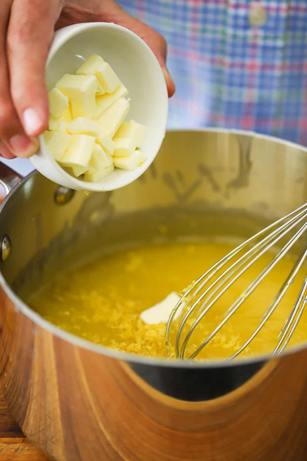 A hand dumping cubed butter from a small white bowl into a metal saucepan filled with lemon curd and lemon zest with a whisk in it.