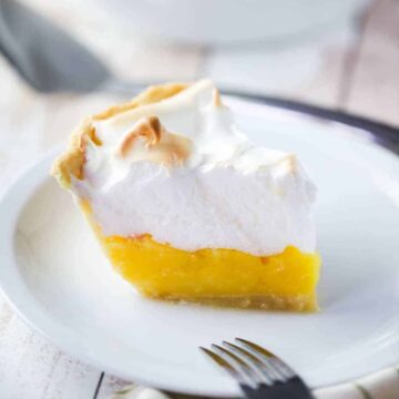 A single slice of a lemon meringue pie on a white plate with the whole pie in the background.