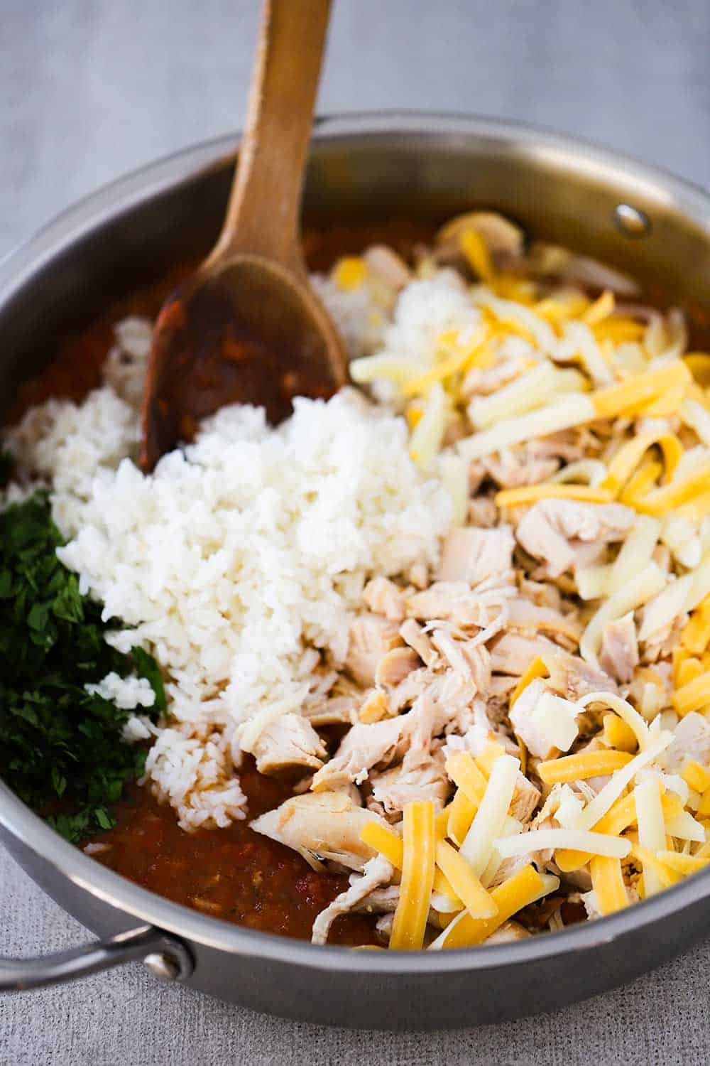 A large silver skillet filled with rice, cooked chicken, and cheese, all being stirred with a wooden spoon into a red sauce.