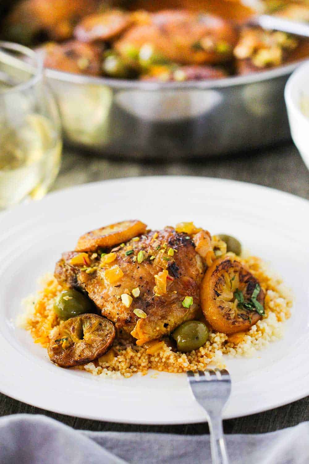 A white dinner plate with a serving of chicken tagine with preserved lemons and olives over a bed of couscous, all sitting next to a skillet filled with the chicken tagine.