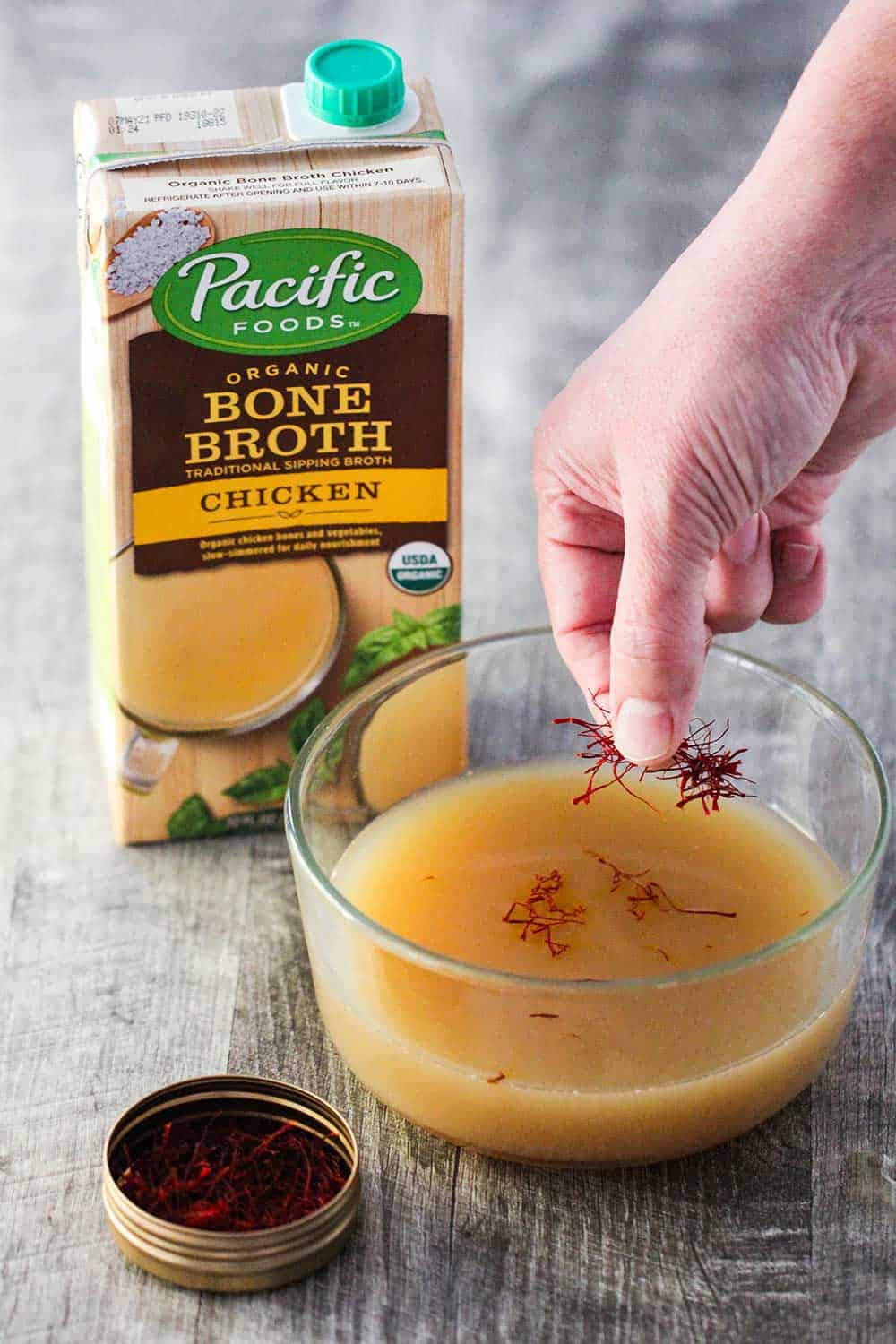 A hand sprinkling saffron threads into a glass bowl of chicken broth next to a box of Pacific Foods chicken broth.
