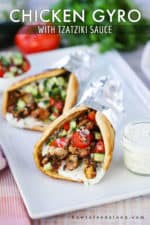 Two chicken gyros wrapped in foil on a white platter next to a small jar filled with tzatziki sauce with a spoon in, with checkered napkins nearby.