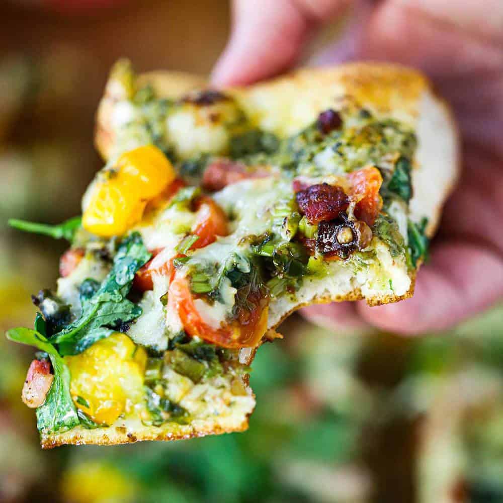 A hand holding a square piece of a BLT Pizza with a bite taken out.