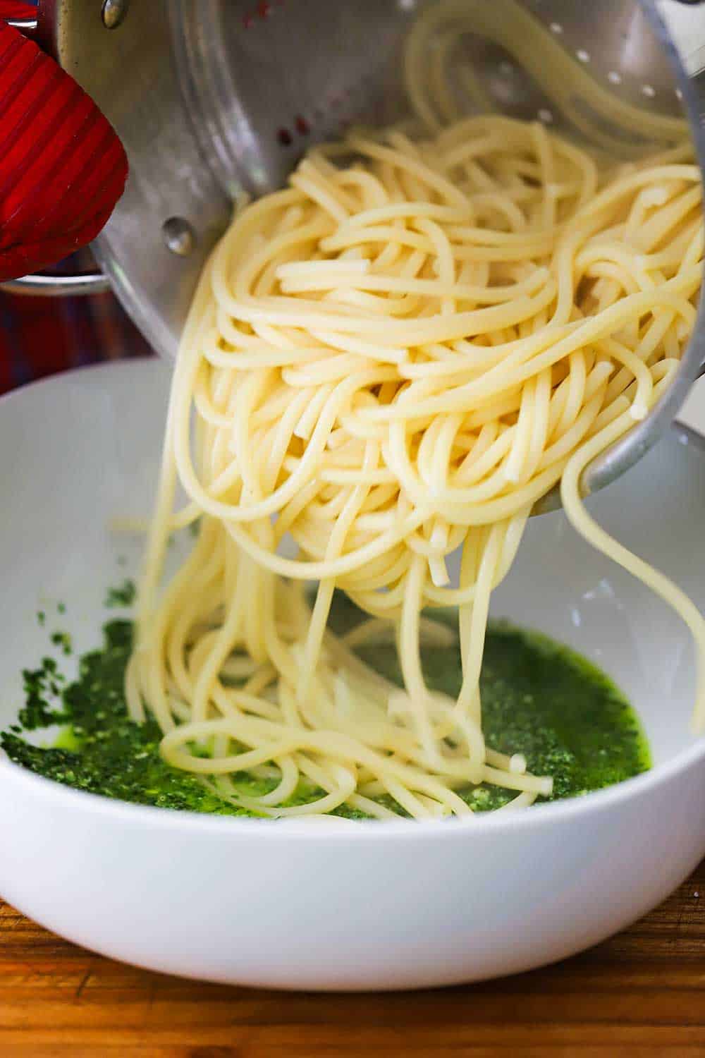 A pasta pot filled cooked spaghetti being transferred into a bowl filled with fresh pesto sauce.