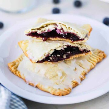 A small white plate holding 2 homemade blueberry pop tarts, one that is split open on top, sitting next to a glass of milk.