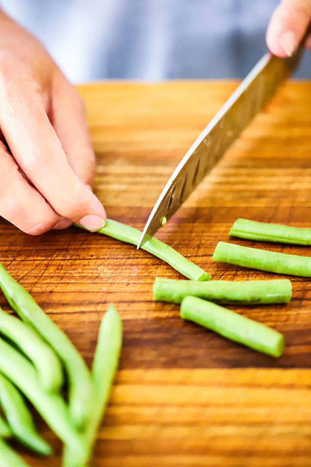 One hand holding a fresh green bean on a cutting board while the other hand uses a large knife to cut it in half.