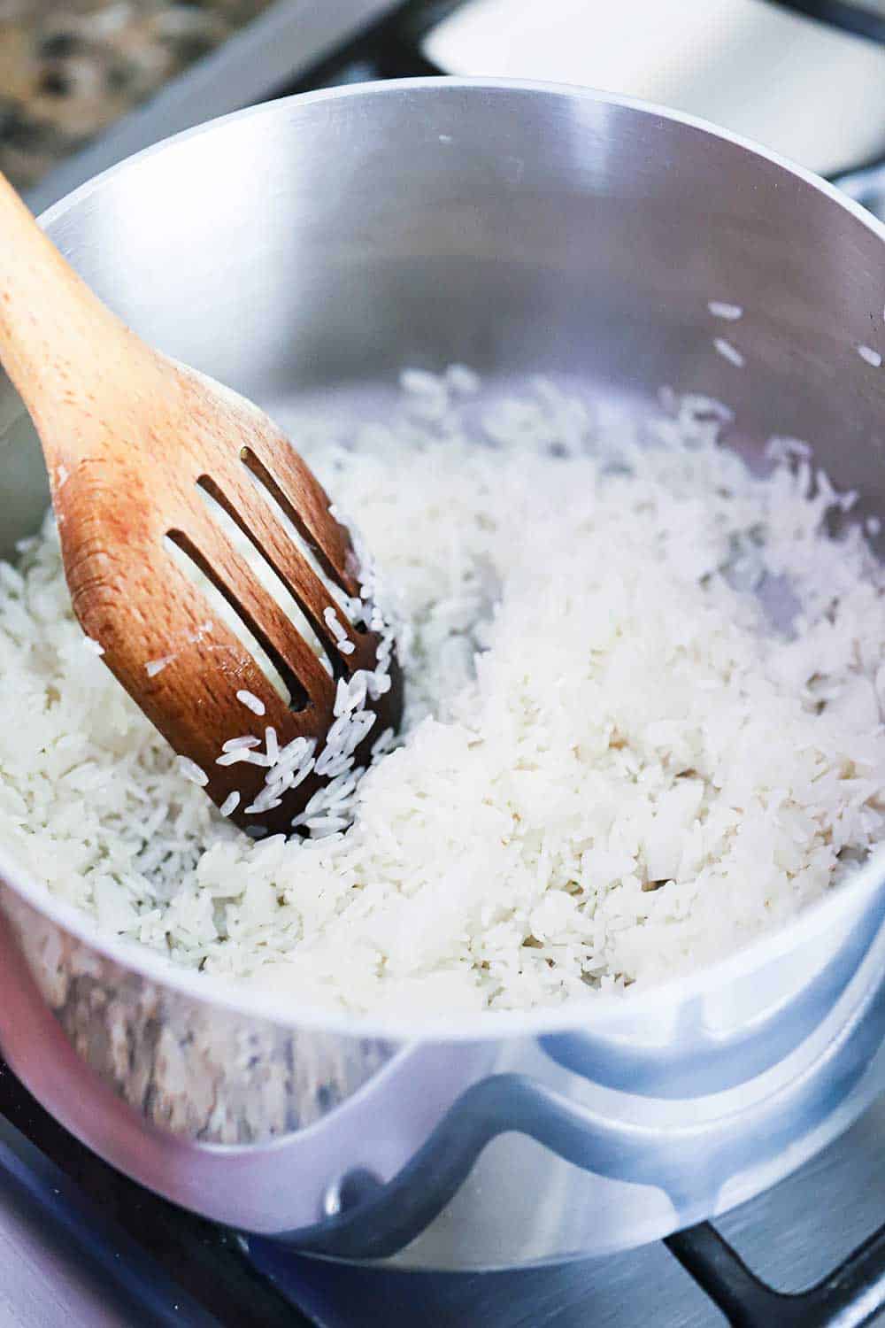 A silver saucepan filled with Jasmine rice and chopped onions being stirred by a slotted wooden spoon.