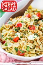 A close-up view of a caprese pasta salad in a large white serving bowl with a wooden spoon in the salad.