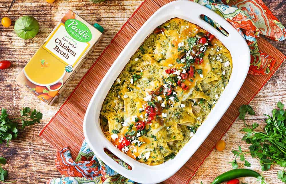 An overhead view of a 9'x13' baking dish filled with chicken enchiladas with verde sauce next to a box of chicken broth and fresh cilantro and cherry tomatoes.