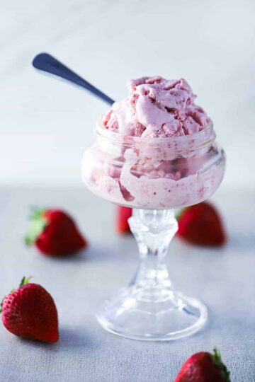A glass ice cream holder filled with homemade strawberry ice cream with a spoon in it and whole strawberries scattered around next to it.