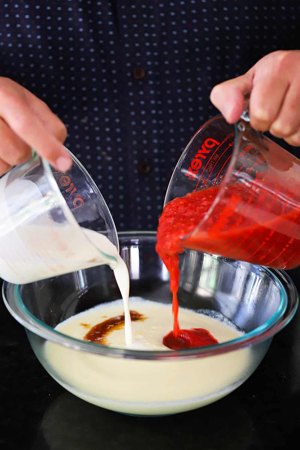 A person using both hands to pour pureed strawberries and heavy cream into a bowl of custard.