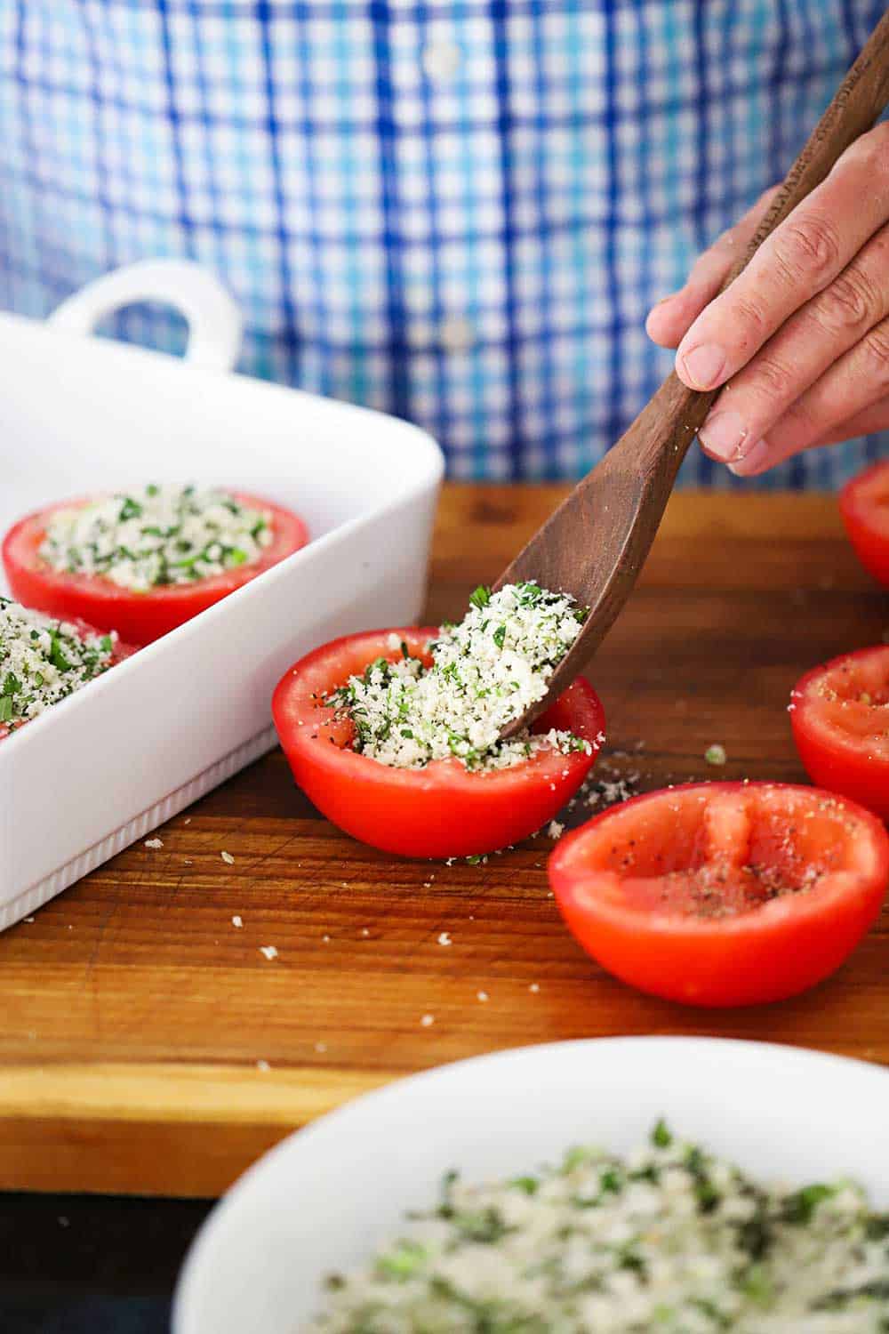 A hand spooning bread crumb filling into the cavity of a tomato half next to a white baking dish.