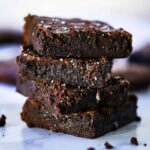 Four stacked fudge brownies on parchment paper with crumbs scattered nearby.