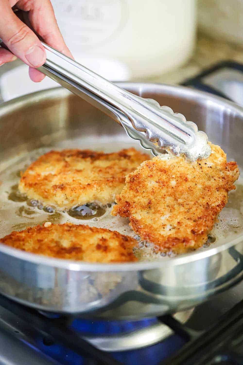 Three chicken cutlets frying in a large saucepan in oil and butter with tongs holding one up.
