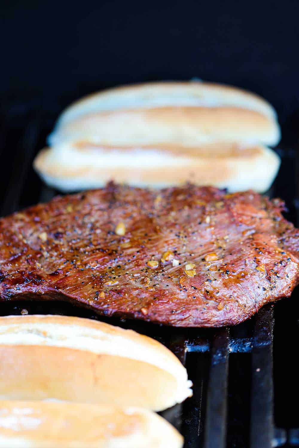A large marinated flank steak on a gas grill with hoagie buns next to it.