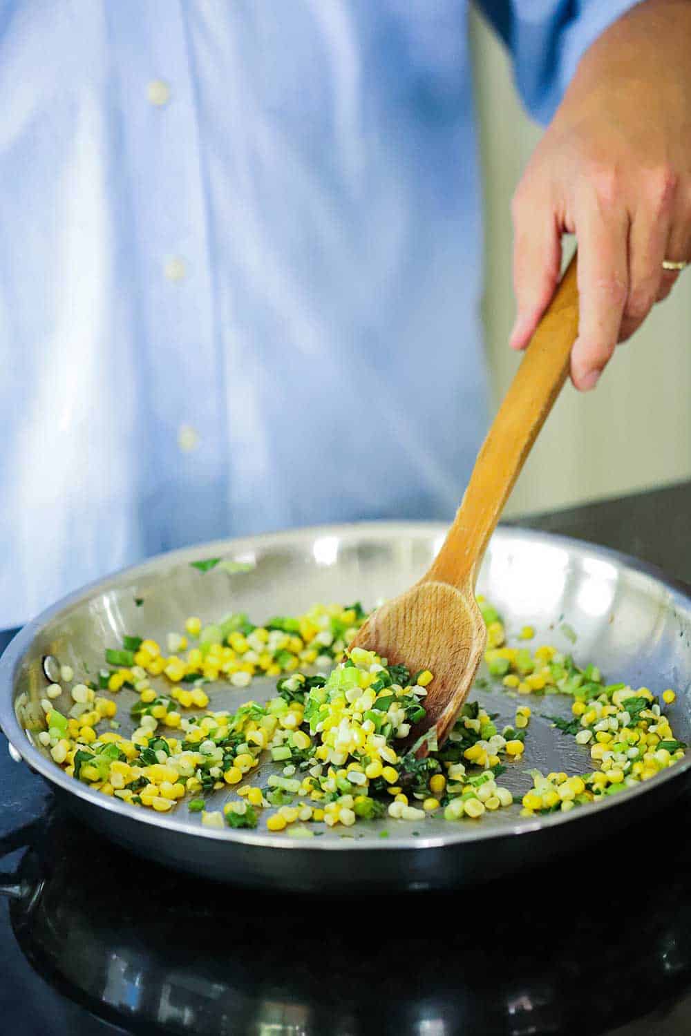 A hand using a wooden spoon to stir corn kernels, sliced scallions, and herbs in a saucepan.