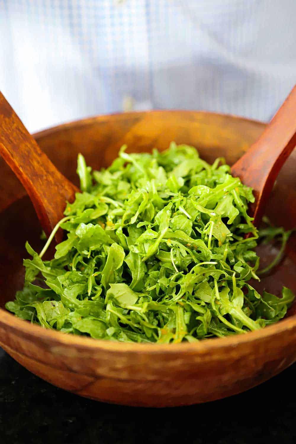 A large wooden salad bowl filled with baby arugula being tossed by a couple of wooden spoons.