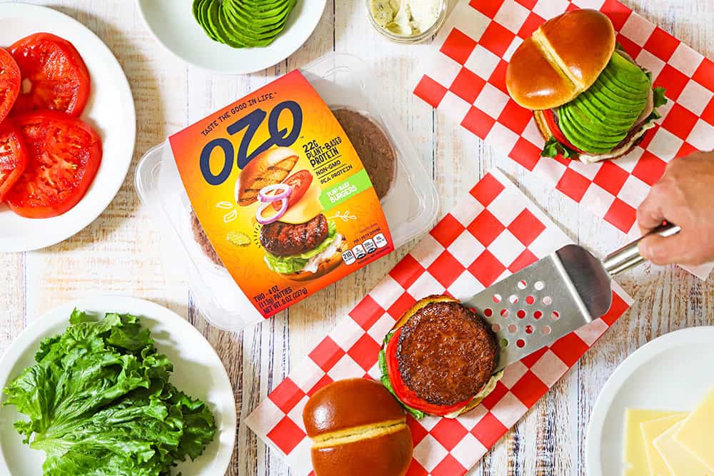 An array of product including sliced tomatoes, leaf lettuce, and a spatula placing a protein-based burger on a bun on a checkered napkin.