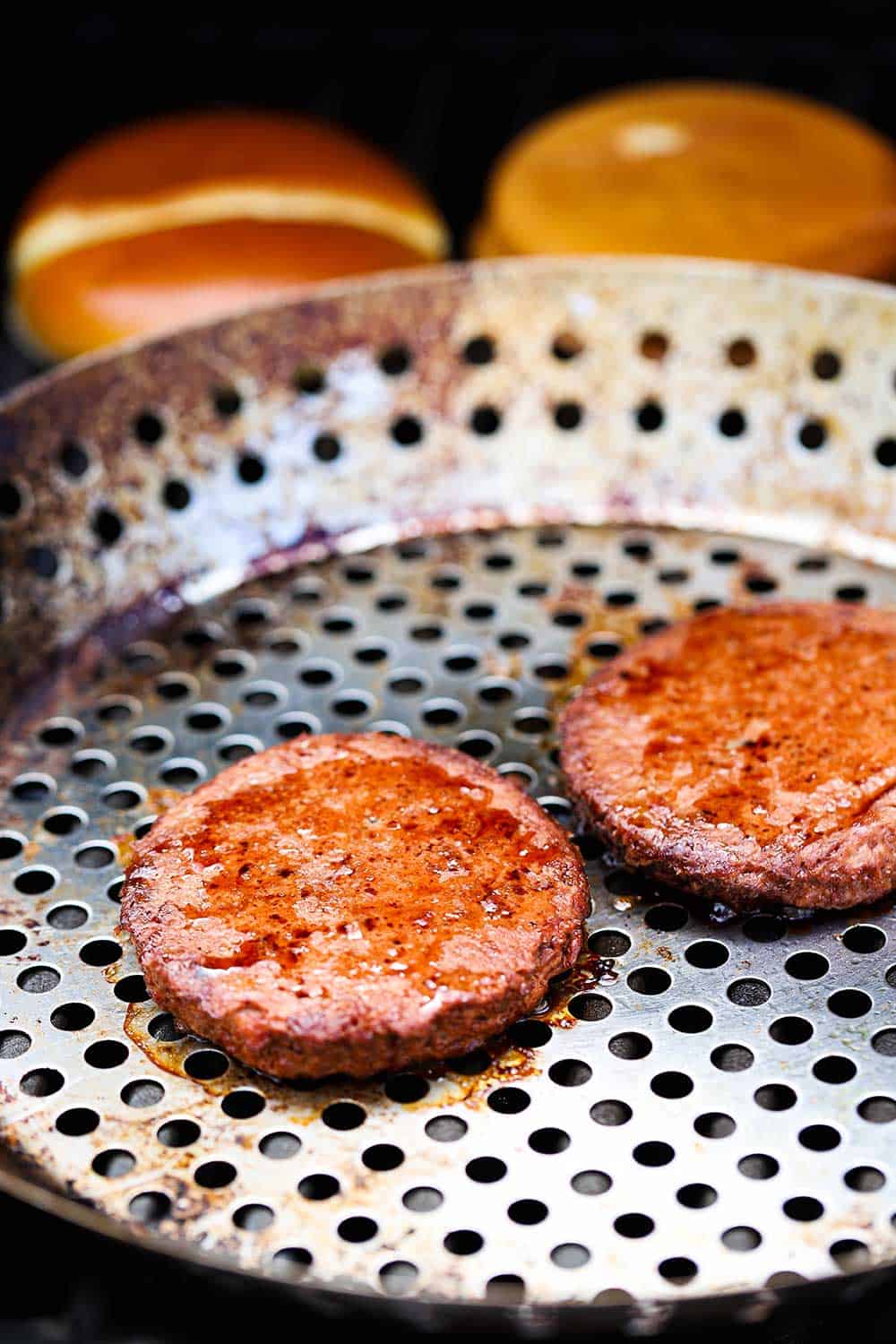 Two plant-based burgers on a grill pan on a grill in front of two hamburger buns, also on the grill.