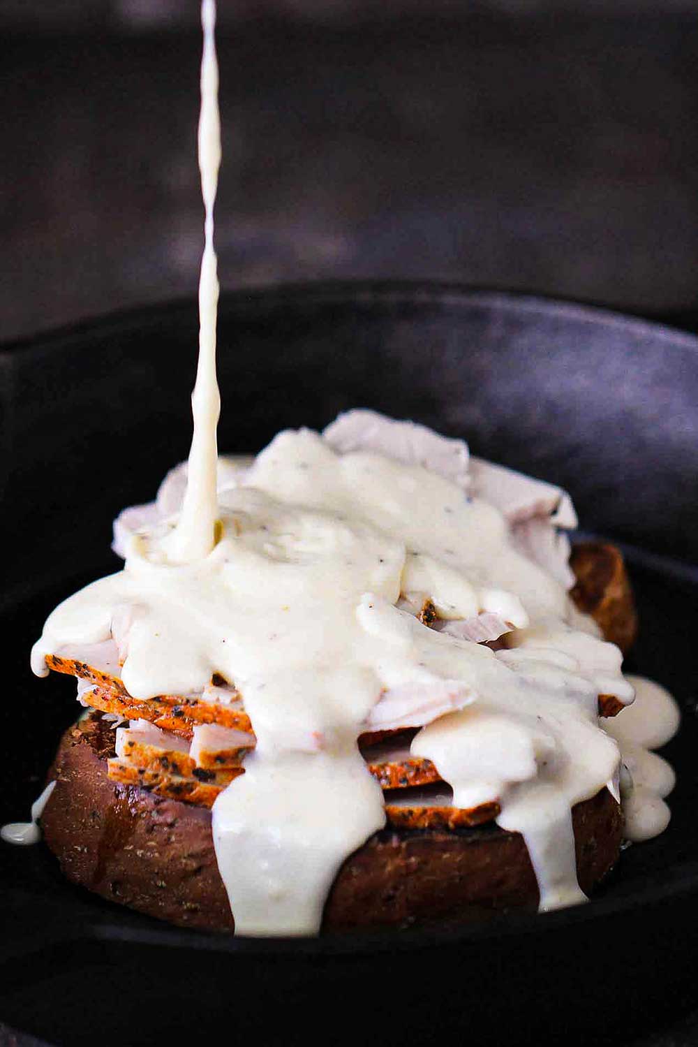 A cast iron skillet filled with a Kentucky hot brown with Mornay sauce being poured over the top.