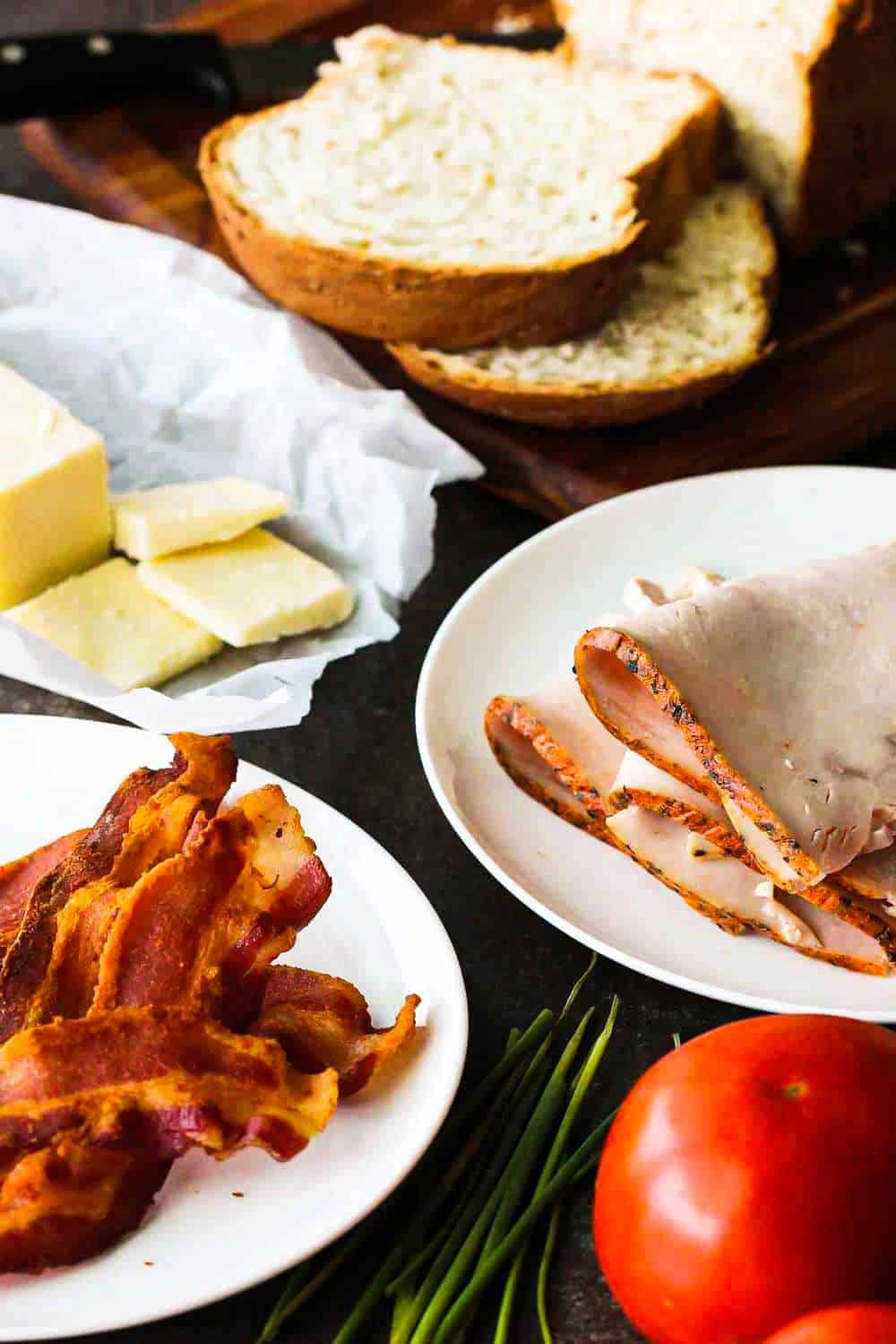 White plates filled with cooked bacon, slice turkey, butter, and surrounded by a tomato and sliced bread.