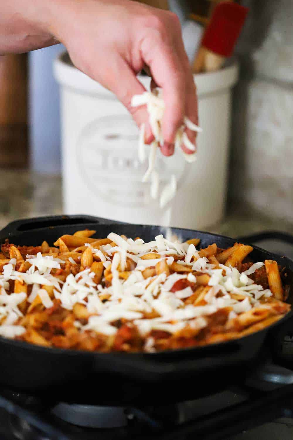 A hand sprinkling grated mozzarella cheese over a pan filled with pasta and sauce.