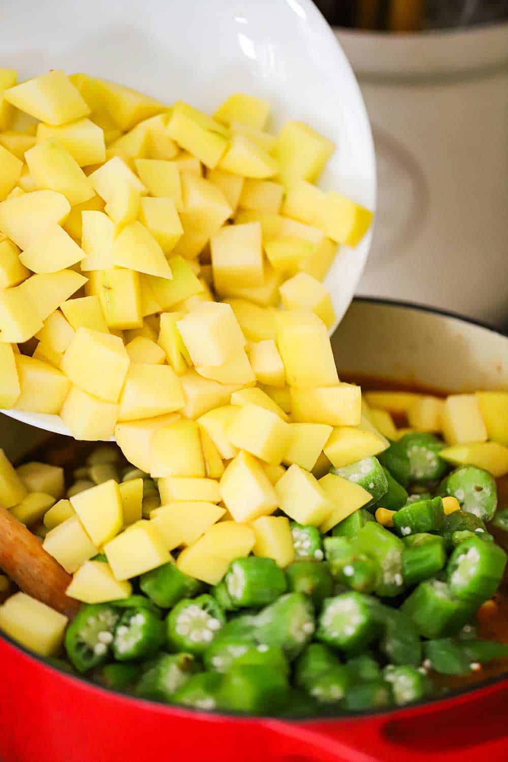 A bowl of cubed yellow potatoes being dumped into a pot of stew topped with sliced okra.