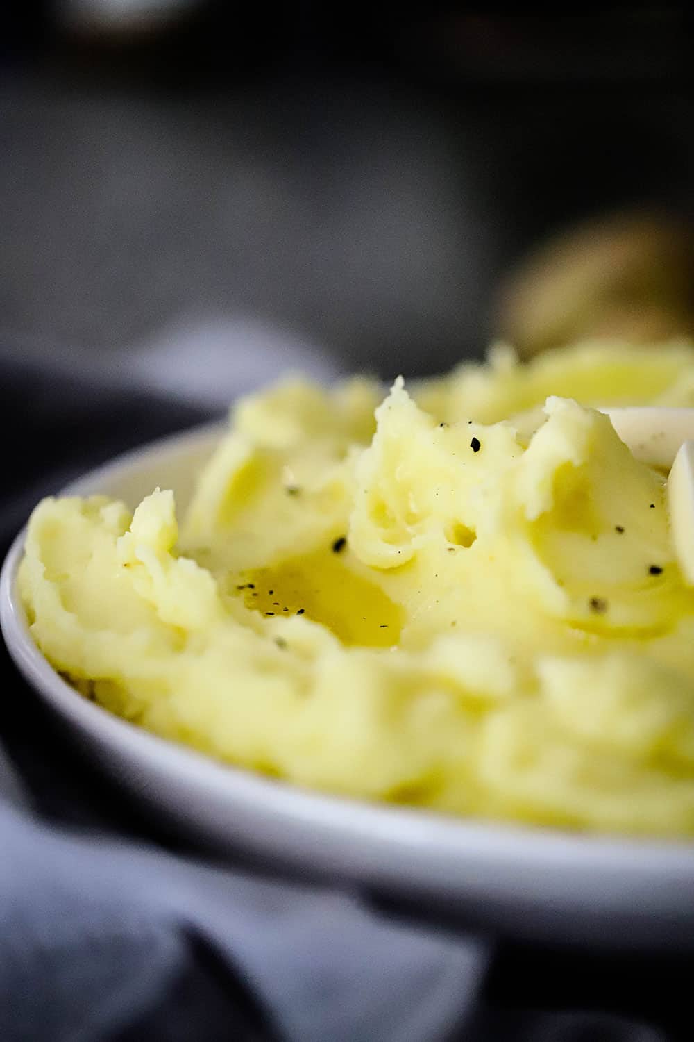 A white serving bowl filled with mashed potatoes with melted butter and ground black pepper on top.