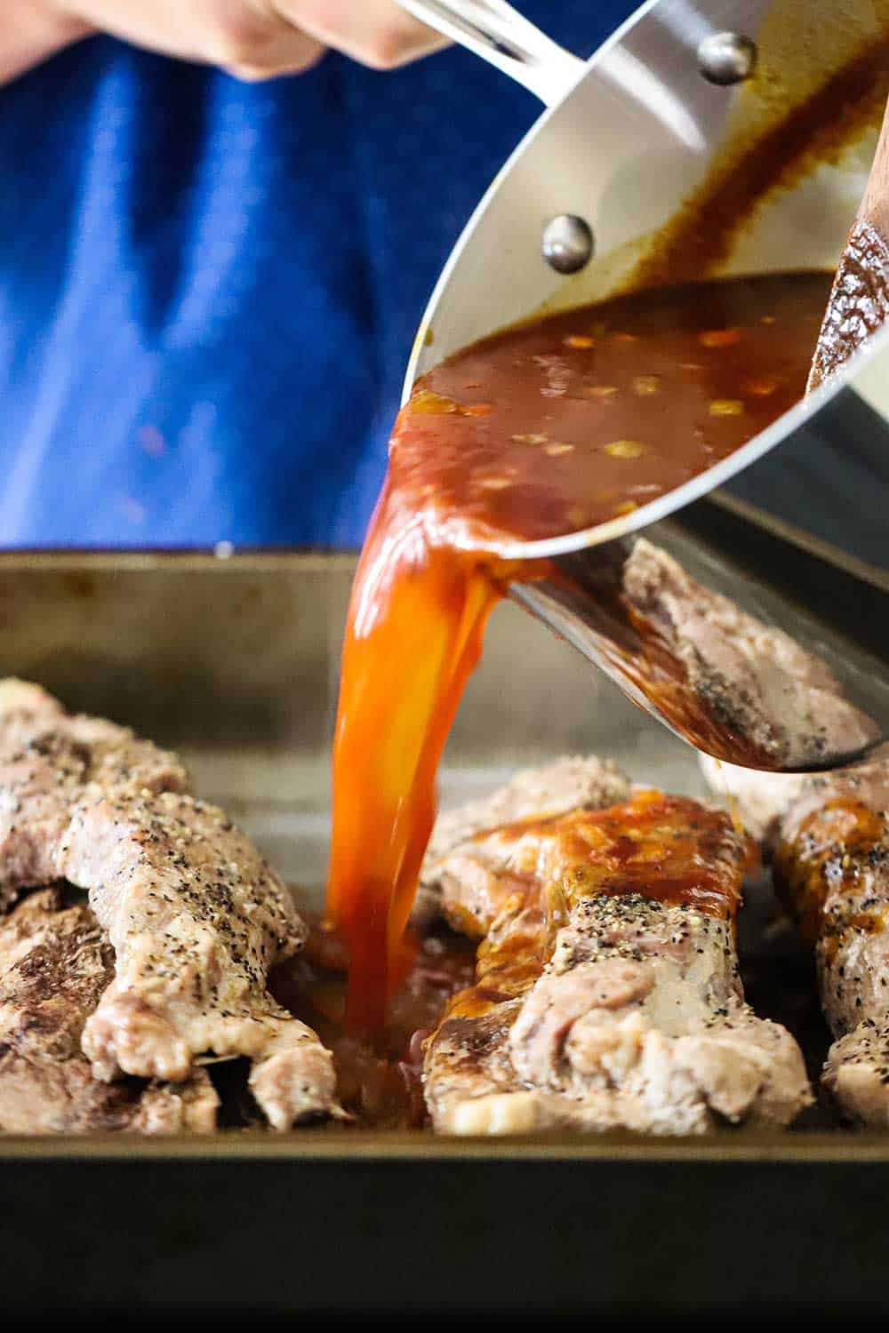 A pan of BBQ sauce being poured over cooked country-style ribs in a large roasting pan.