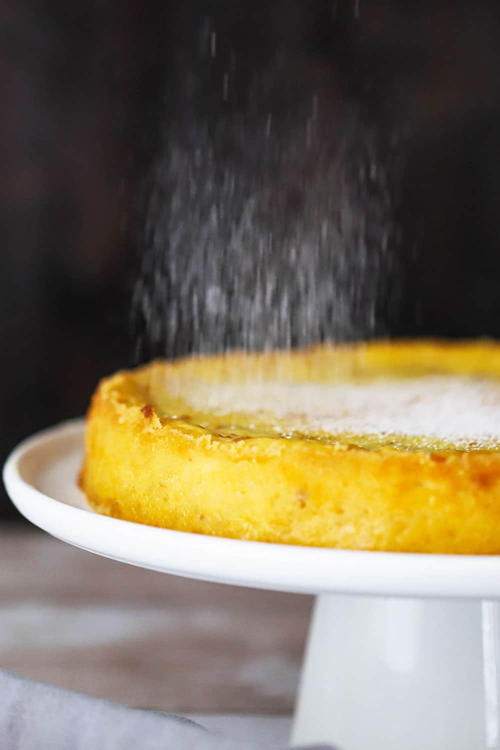Powdered sugar sprinkling over the top of Italian ricotta cheesecake on a cake stand.