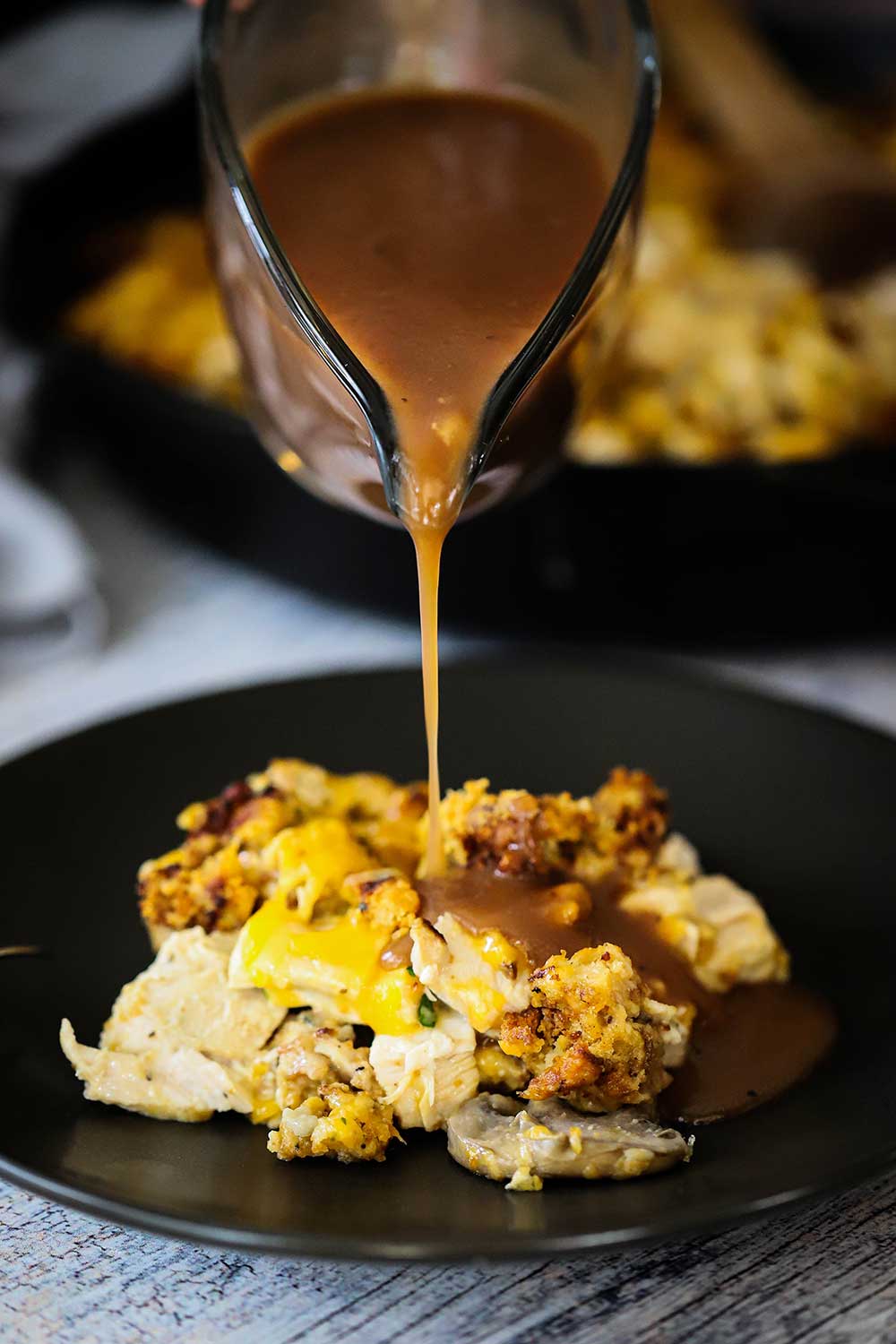 Brown gravy being poured from a glass gravy boat onto a plate of turkey and stuffing casserole.